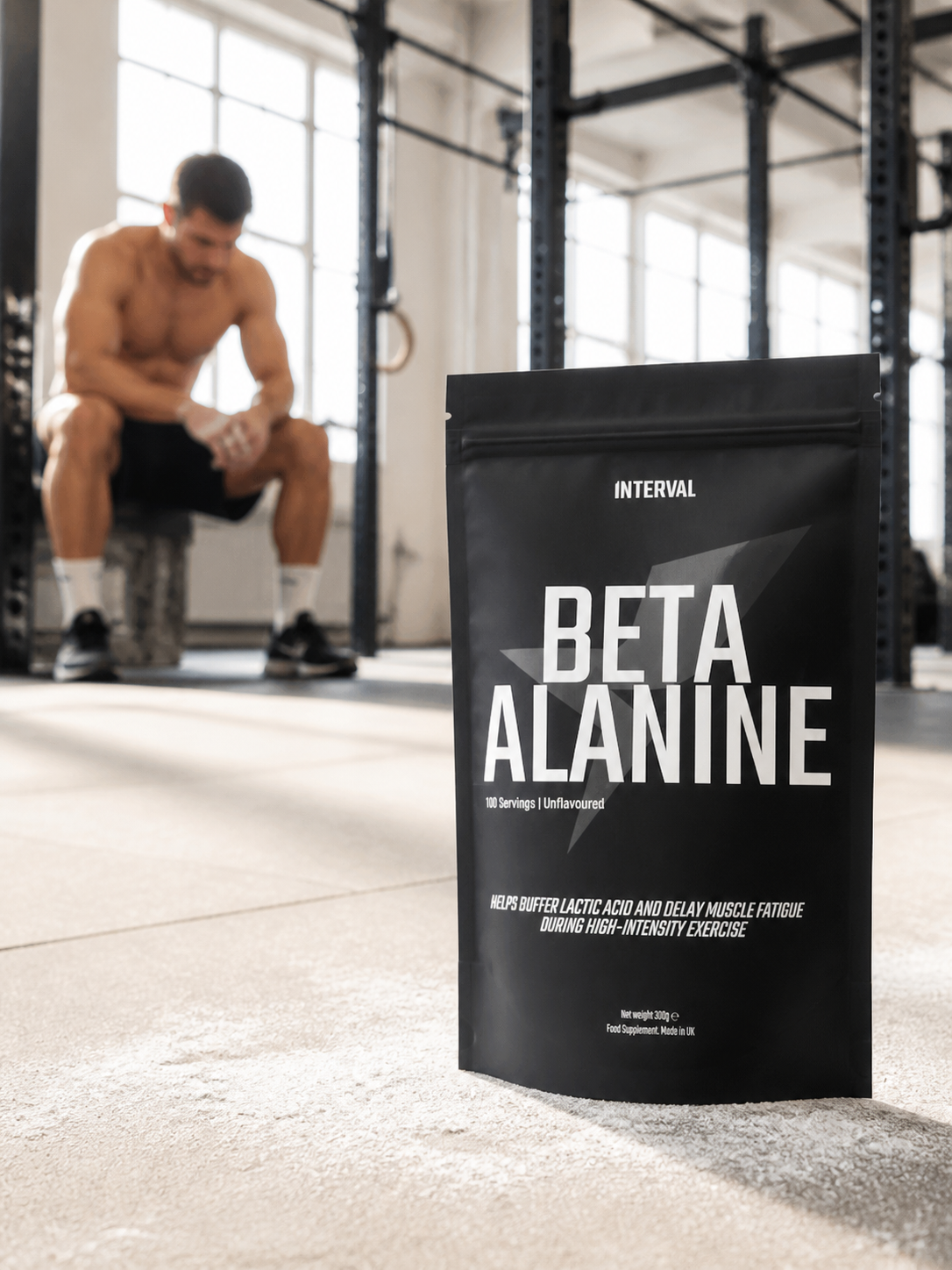 A black stand-up pouch of INTERVAL Beta Alanine powder stands on a gym floor in the foreground, with a blurred shirtless man sitting on a bench in a gym in the background.