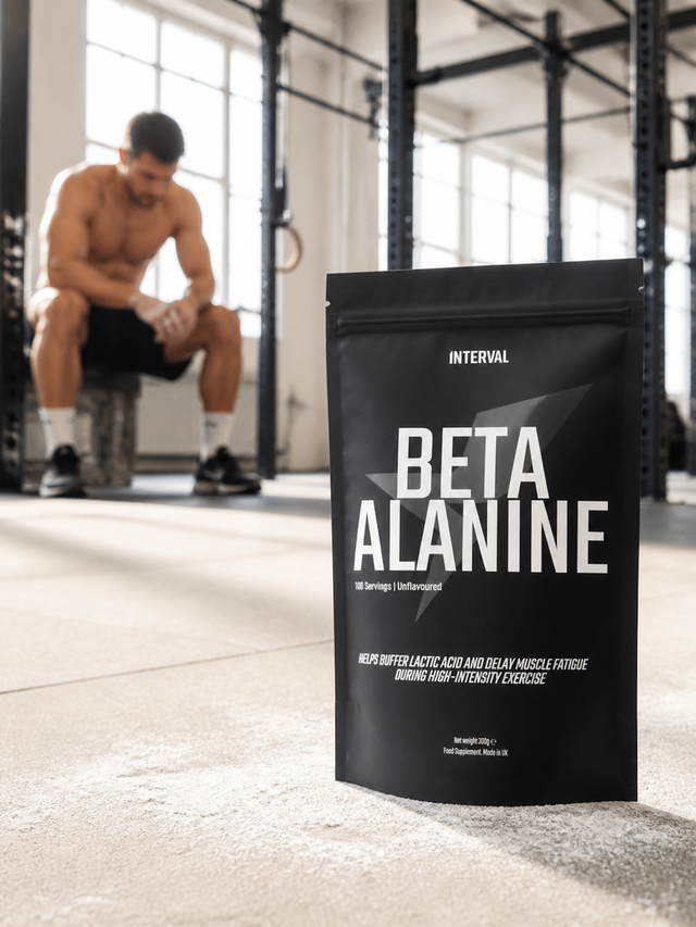 A black stand-up pouch of INTERVAL Beta Alanine powder stands on a gym floor in the foreground, with a blurred shirtless man sitting on a bench in a gym in the background.
