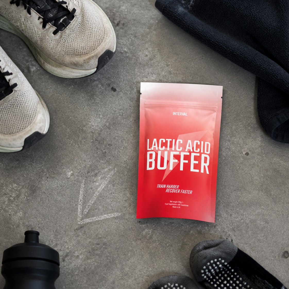 A red and white pouch of Lactic Acid Buffer electrolyte drink mix lies on a gray concrete floor, surrounded by athletic gear including a water bottle, running shoes, a black jacket, and grip socks.