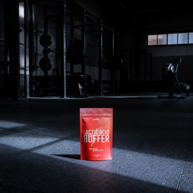 A red stand-up pouch labeled 'LACTIC ACID BUFFER' stands on a gym floor in dramatic low light, with exercise equipment blurred in the background.