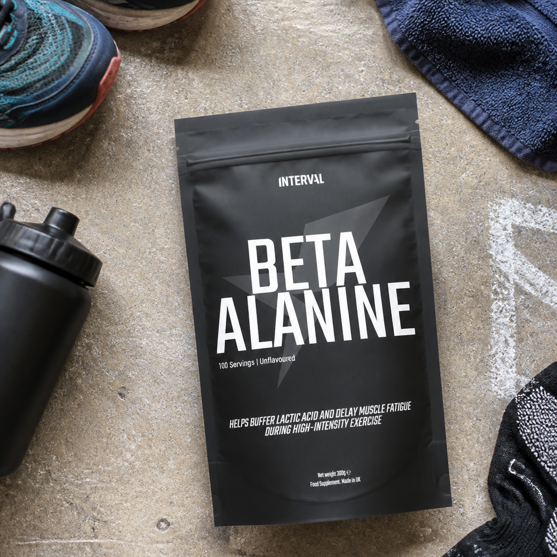 A black stand-up pouch of INTERVAL Beta Alanine supplement placed on a concrete floor, surrounded by athletic shoes, a towel, a water bottle, and socks.