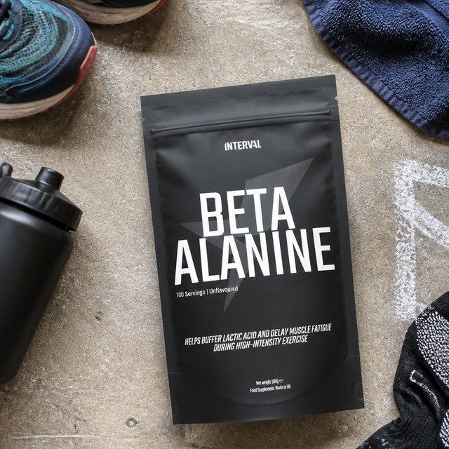 A black stand-up pouch of INTERVAL Beta Alanine supplement placed on a concrete floor, surrounded by athletic shoes, a towel, a water bottle, and socks.