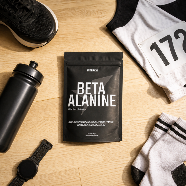 A black stand-up pouch labeled INTERVAL BETA ALANINE sits on a light wood surface, surrounded by a black water bottle, a black watch, a black athletic shoe, a white jersey with the number 172, and white-and-black socks.